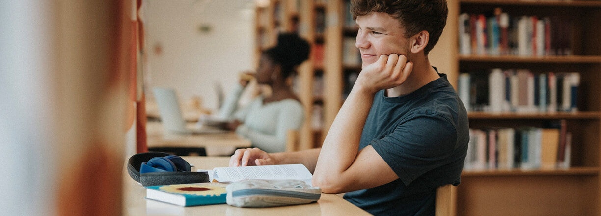 Um estudante estudando em uma mesa com livros e um laptop em uma biblioteca.