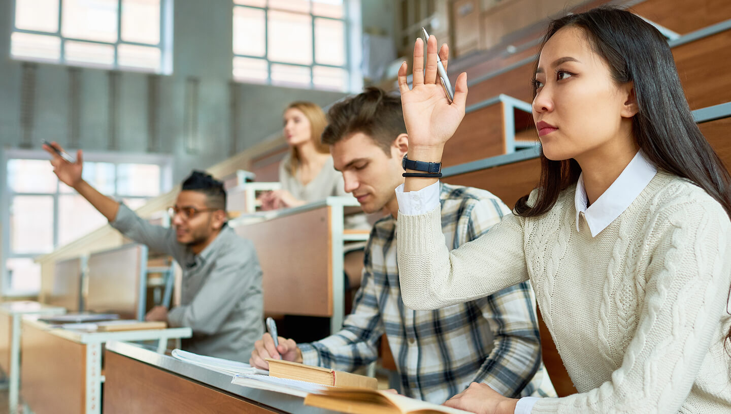 Uma sala de aula universitária com estudantes sentados em mesas, enquanto uma estudante em primeiro plano levanta a mão.