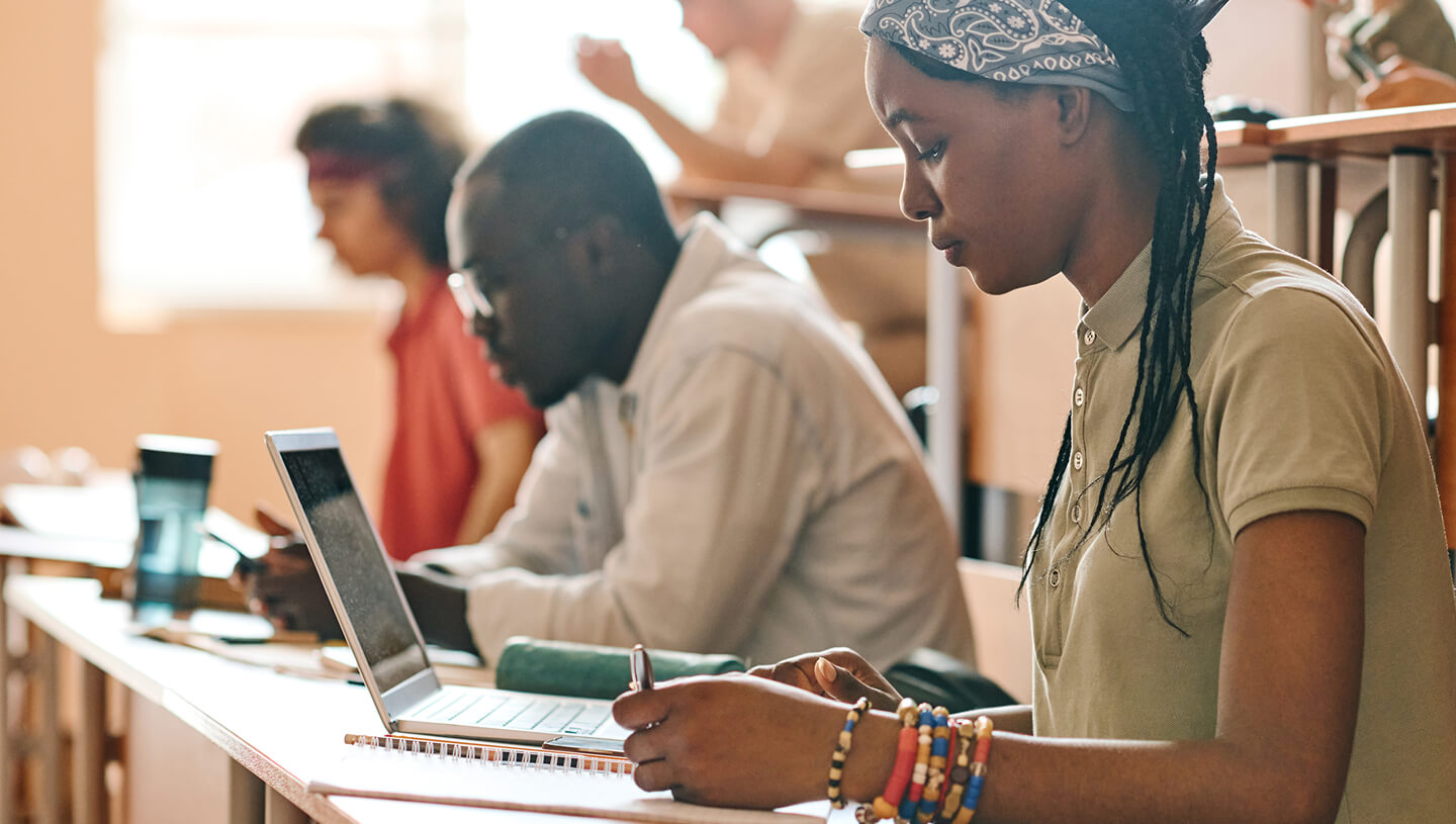 Alunos trabalhando em uma sala de aula, com um aluno usando um laptop e fazendo anotações em uma mesa.